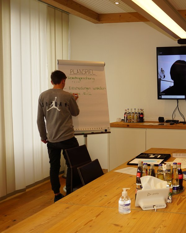 Junge, der etwas auf ein Flipchart schreibt, weiterer junge der an einem Besprechungstisch sitzt und zwei Personen die über ein Teamsmeeting zugeschalten sind | © RÖSLE GROUP