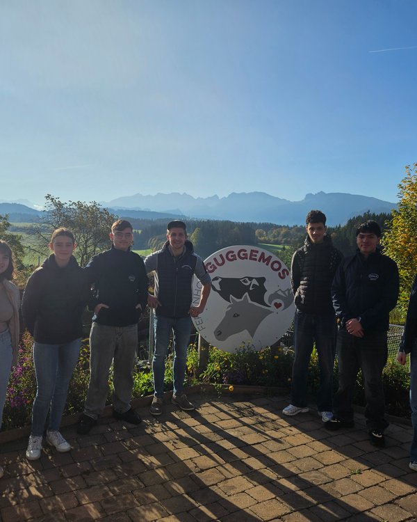 Auf dem Bild stehen 7 junge Menschen um ein Schild auf dem "Ferienhof Guggemos" steht. Alle lächeln in die Kamera | © RÖSLE GROUP