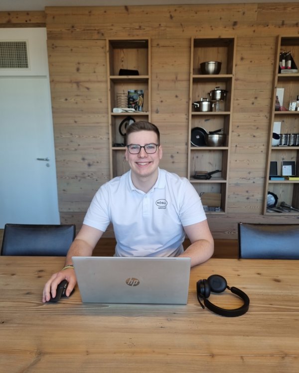 Ein junger Mann sitzt an einem Holztisch. vor ihm ist ein Laptop aufgeschlagen und daneben ein Headset. Er lächelt in die Kamera | © RÖSLE GROUP